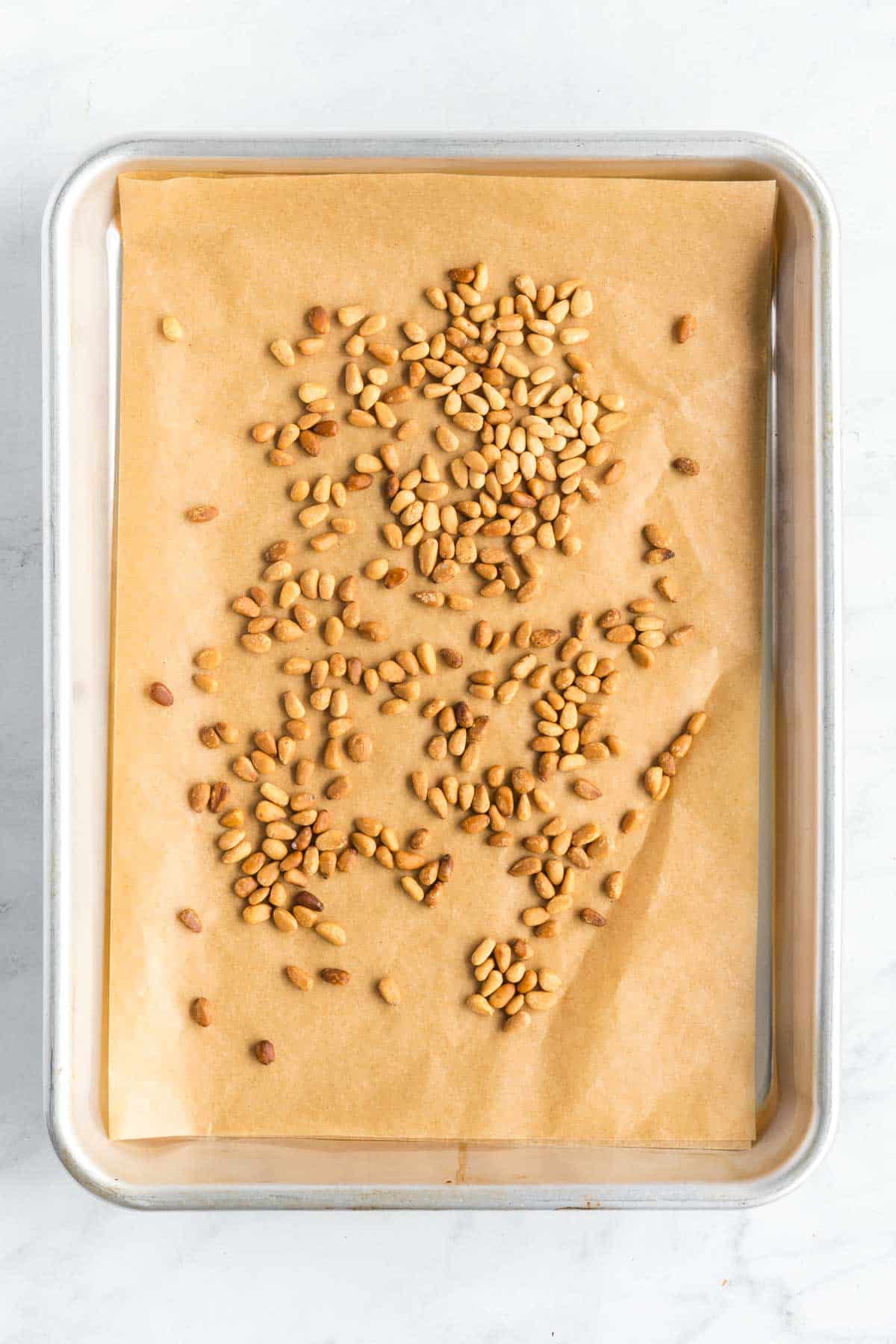 A baking sheet lined with parchment paper holds an uneven layer of toasted pine nuts.