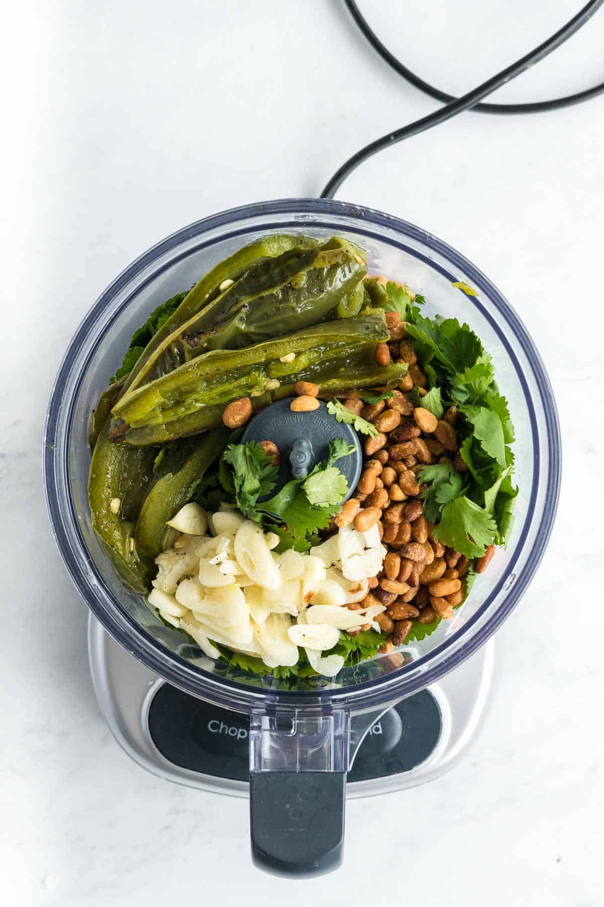 Food processor filled with roasted green chilies, garlic cloves, peanuts, and cilantro, viewed from above on a white surface.