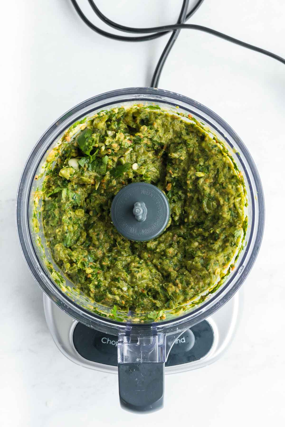 A food processor filled with a blended green mixture, possibly a sauce or dip, sits on a white countertop.