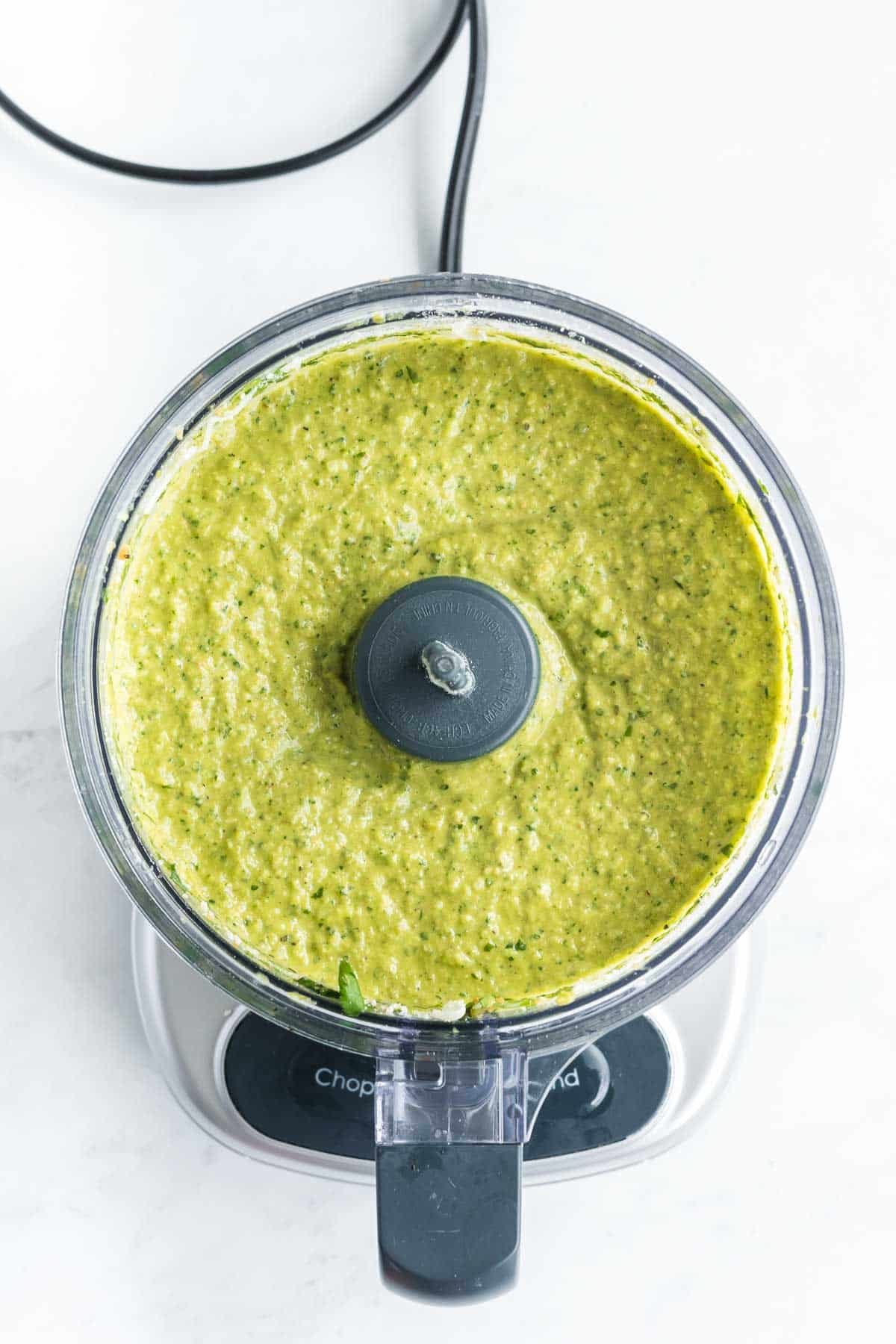 Overhead view of a food processor filled with a blended green sauce on a white surface.