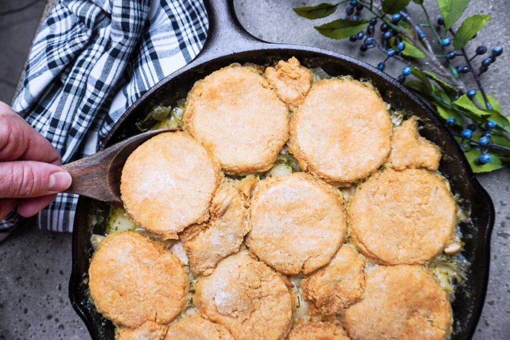 A hand scoops biscuit-topped savory pie from a cast iron skillet; a plaid towel and a sprig with blue berries are nearby.