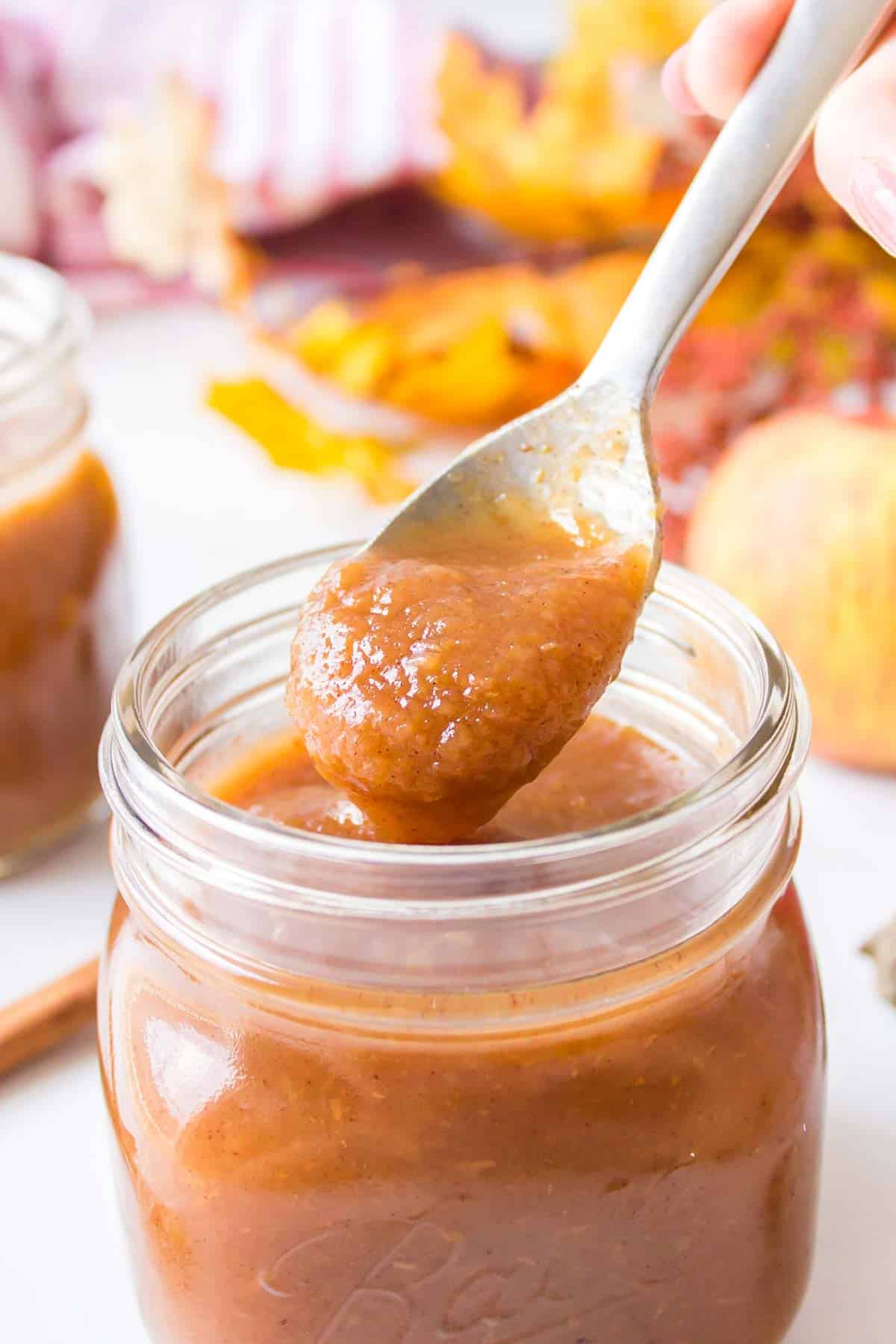 A spoon scooping thick, brown apple butter from a glass jar, with autumn leaves and another jar in the background.