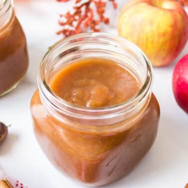 Two jars of apple butter on a white surface, surrounded by fresh apples and autumn decorations.