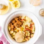 A white slow cooker filled with chopped apples, brown sugar, and spices. Surrounding are lemon halves, a measuring cup with liquid, an empty small jar, and a pink-striped towel.