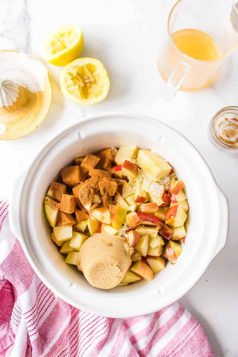 A white slow cooker filled with chopped apples, brown sugar, and spices. Surrounding are lemon halves, a measuring cup with liquid, an empty small jar, and a pink-striped towel.