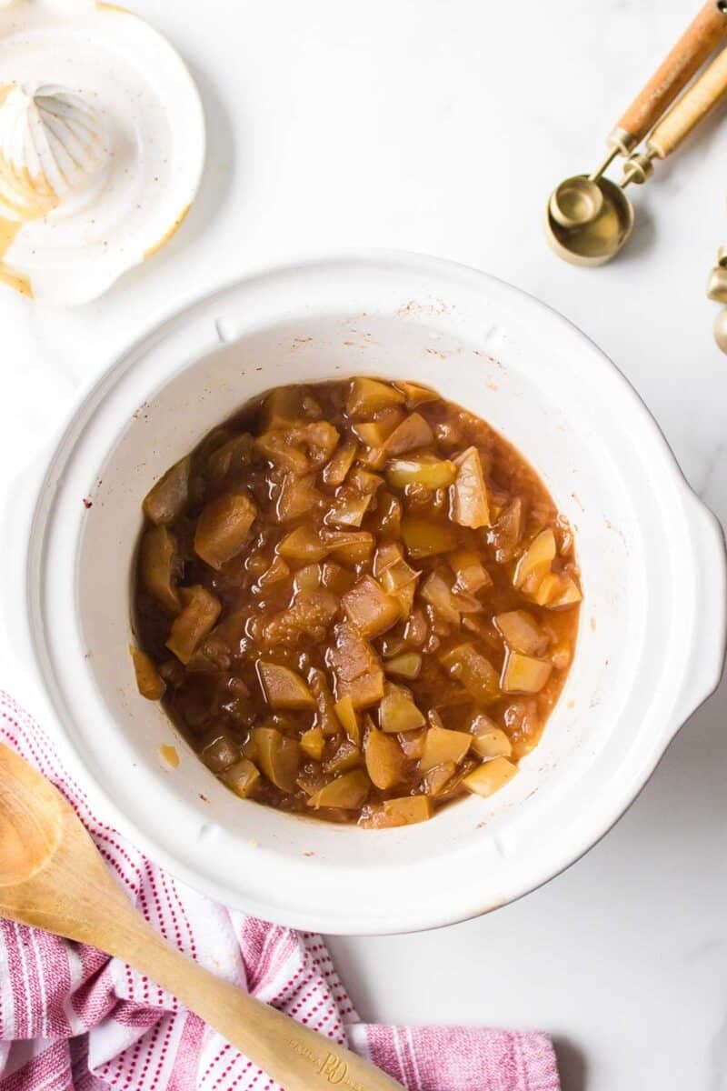 A white slow cooker filled with cooked, chunky apple filling sits on a white surface next to a wooden spoon, a red-striped towel, and measuring spoons.