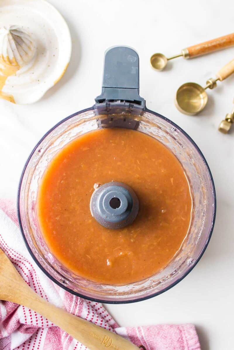 Food processor bowl filled with a thick, light brown sauce, surrounded by measuring spoons, a citrus juicer, a dish towel, and a wooden spoon on a white surface.