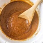 A wooden spoon rests in a white slow cooker filled with thick, brown apple butter.
