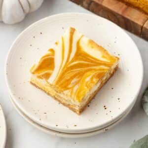 A square piece of pumpkin cheesecake bar with a swirled orange and white pattern on a white speckled plate.