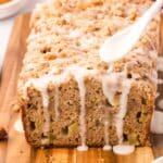 A loaf of apple streusel bread on a wooden board is being drizzled with white icing from a spoon.