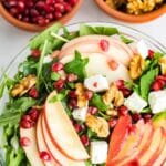 A bowl of salad with arugula, apple slices, walnuts, pomegranate seeds, and cubes of feta cheese. Two small bowls with pomegranate seeds and walnuts are in the background.