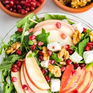 A bowl of salad with arugula, apple slices, walnuts, pomegranate seeds, and cubes of feta cheese. Two small bowls with pomegranate seeds and walnuts are in the background.