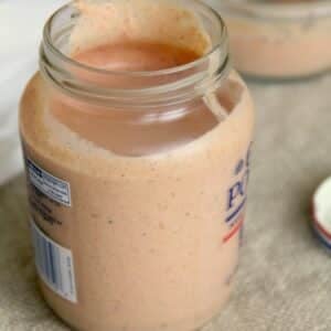 An open glass jar filled with a creamy, light orange sauce or dressing sits on a textured surface next to a jar lid.