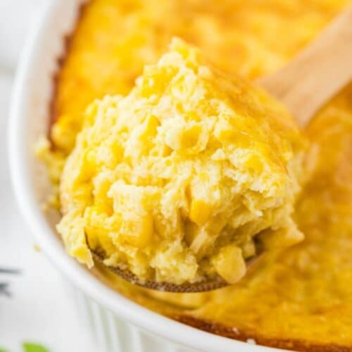 A close-up of a wooden spoon scooping out a portion of baked corn casserole from a white dish.