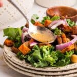 A spoon drizzles dressing over a salad with kale, roasted sweet potatoes, red onion, and chickpeas on a stacked plate.