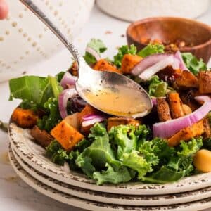 A spoon drizzles dressing over a salad with kale, roasted sweet potatoes, red onion, and chickpeas on a stacked plate.