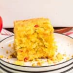 A slice of cornbread with visible pieces of red and green peppers sits on a white plate with a black rim. Crumbs are scattered on the plate.