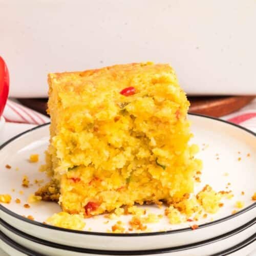 A slice of cornbread with visible pieces of red and green peppers sits on a white plate with a black rim. Crumbs are scattered on the plate.
