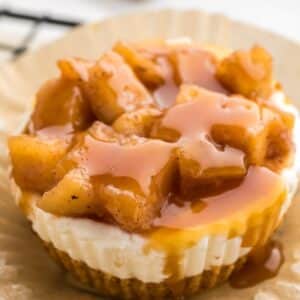 Mini mini caramel apple cheesecake topped with spiced apple chunks and drizzled with caramel sauce, resting on a brown paper liner.