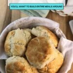 A basket lined with a cloth holds four golden, homemade biscuits—perfect for adding to your favorite meals; a plate and utensils are partially visible in the background.