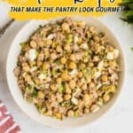 A bowl of tuna salad with chickpeas, celery, and eggs is on a white surface with a red striped towel nearby. Text reads, "11 Tuna Recipes That Make the Pantry Look Gourmet.