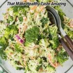A glass bowl filled with broccoli salad mixed with shredded carrots, red onions, and dressing, with serving utensils inside the bowl.