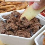 A hand dips an apple slice into a bowl of chocolate chip dessert dip, with pretzels and a spoon nearby.