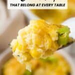 A spoonful of creamy corn casserole garnished with chopped green onions, with more casserole blurred in the background. Text reads: "14 Corn Recipes That Belong at Every Table.