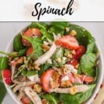 A bowl of salad containing spinach, shredded chicken, sliced strawberries, walnuts, and crumbled cheese on a white surface.
