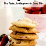 A stack of strawberry cookies on a white plate with a sliced strawberry beside it; blurred background features a bowl of strawberries and a milk bottle.