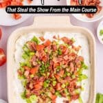 A creamy dip topped with chopped lettuce, tomatoes, bacon, and green onions in a square dish, surrounded by plates and a bowl of chopped green onions.