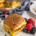 A breakfast sandwich with egg and bacon on a plate, garnished with herbs, served with blackberries and raspberries; more sandwiches in the background.