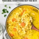 A pot of chicken noodle soup with carrots and herbs sits on a white surface next to chopped parsley and a striped napkin.