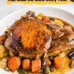 A cooked pork chop sits on a white plate surrounded by roasted carrots, potatoes, and green beans, with a brown sauce.