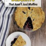A whole blueberry pie with a slice cut out on a small white plate beside it, placed on a wooden surface with a striped kitchen towel nearby—perfect for fans of classic blueberry desserts.