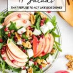 A bowl of salad with apple slices, feta cheese, arugula, pomegranate seeds, and walnuts on a table, with walnuts and an apple nearby.