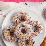 A plate of donuts with white icing and orange, purple, and black Halloween-themed sprinkles, surrounded by a glass of milk and festive decorations.
