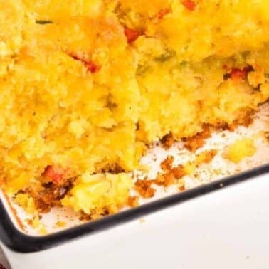 A close-up of a partially eaten cornbread casserole with a golden crust, showing a moist, crumbly interior with pieces of red and green peppers.