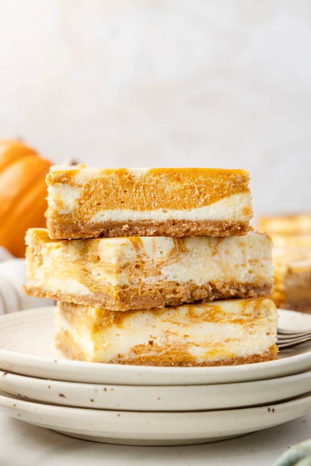 Three pumpkin swirl cheesecake bars are stacked on a plate, showing layers of creamy cheesecake and pumpkin filling on a crumbly crust.