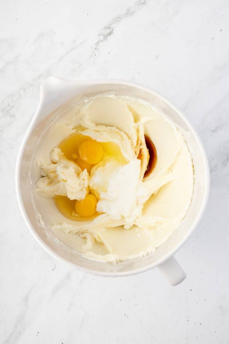 A mixing bowl containing creamed butter and sugar with two cracked eggs and vanilla extract on top, ready to be mixed.
