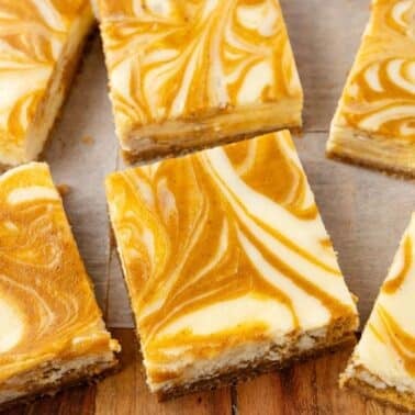 Six pumpkin swirl cheesecake bars are arranged on parchment paper over a wooden surface. The bars have a marbled orange and cream pattern on top.
