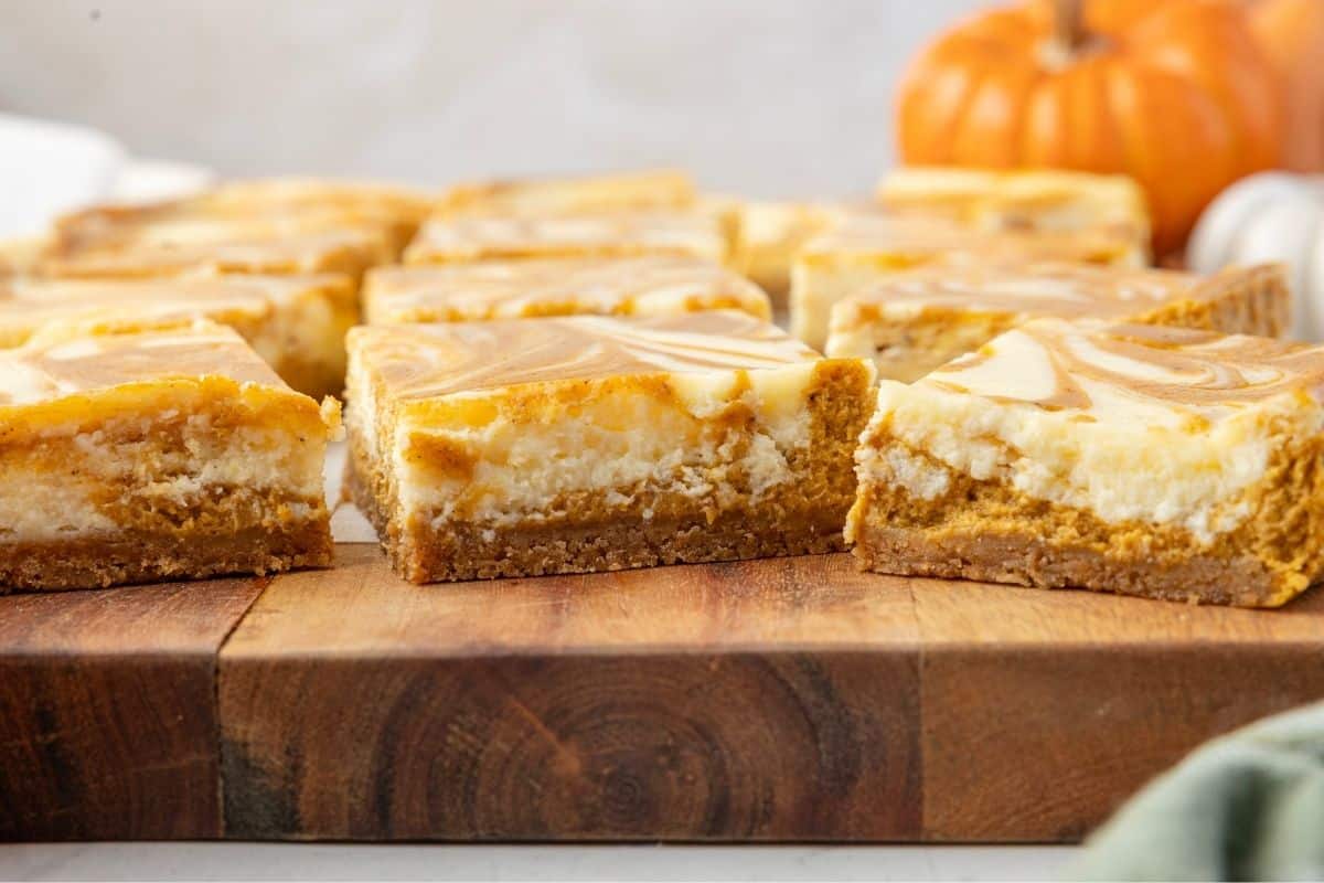 Several pumpkin cheesecake bars with a swirled top are arranged on a wooden cutting board, with a small pumpkin visible in the background.