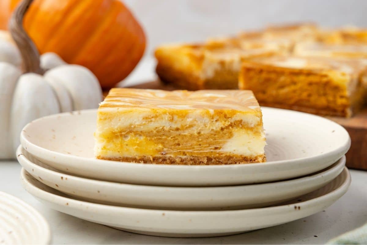 A square piece of marbled pumpkin cheesecake sits on a stack of white plates, with small pumpkins and more cheesecake pieces in the background.