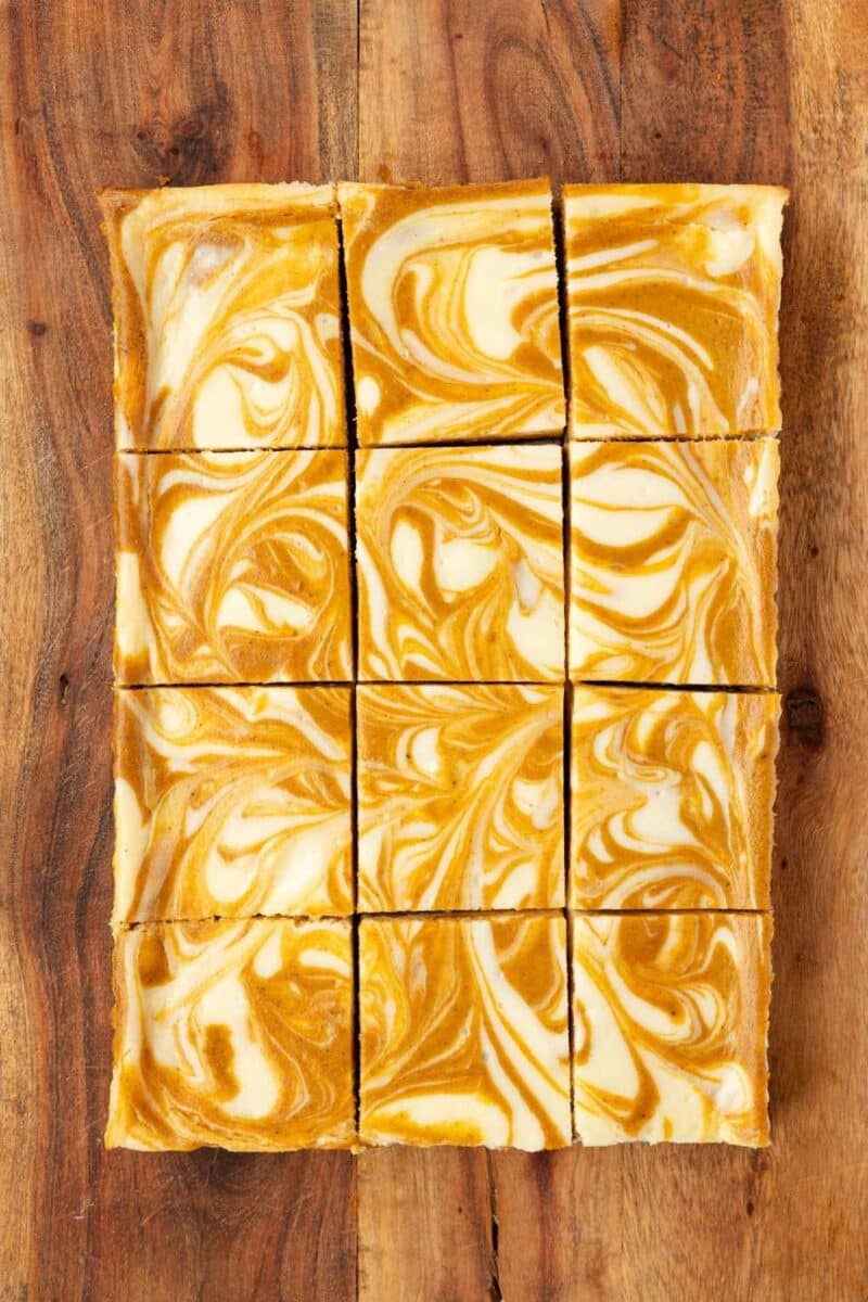 Nine pumpkin swirl cheesecake bars arranged in a rectangle on a wooden surface, featuring a marbled orange and white pattern on top.