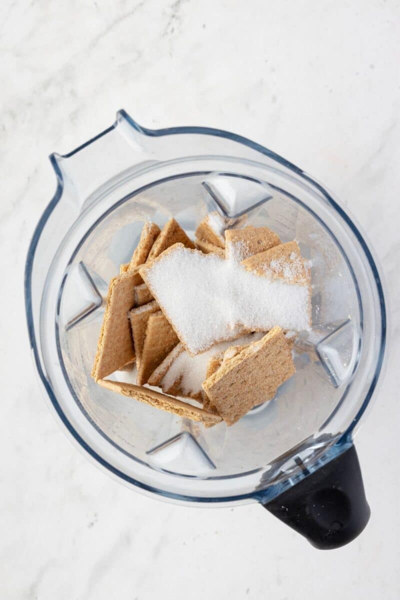 A blender pitcher containing broken graham crackers and a pile of granulated sugar on a white surface.