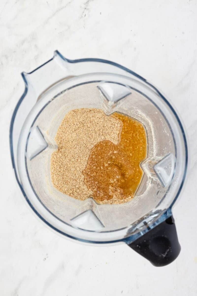 Overhead view of a blender containing ground almonds and a brown liquid mixture on a white surface.