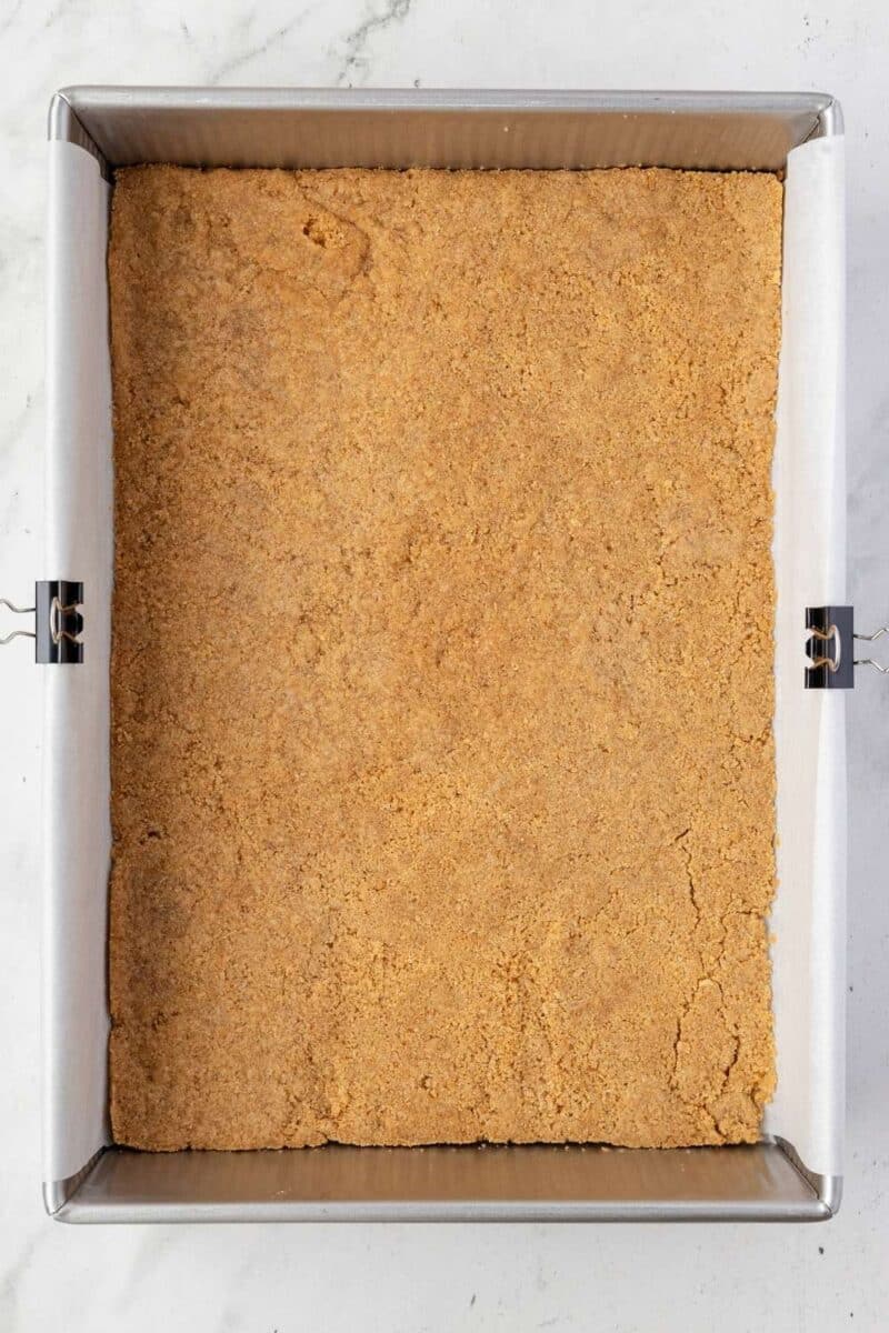 A square baking pan lined with parchment paper holds an even layer of pressed graham cracker crust.