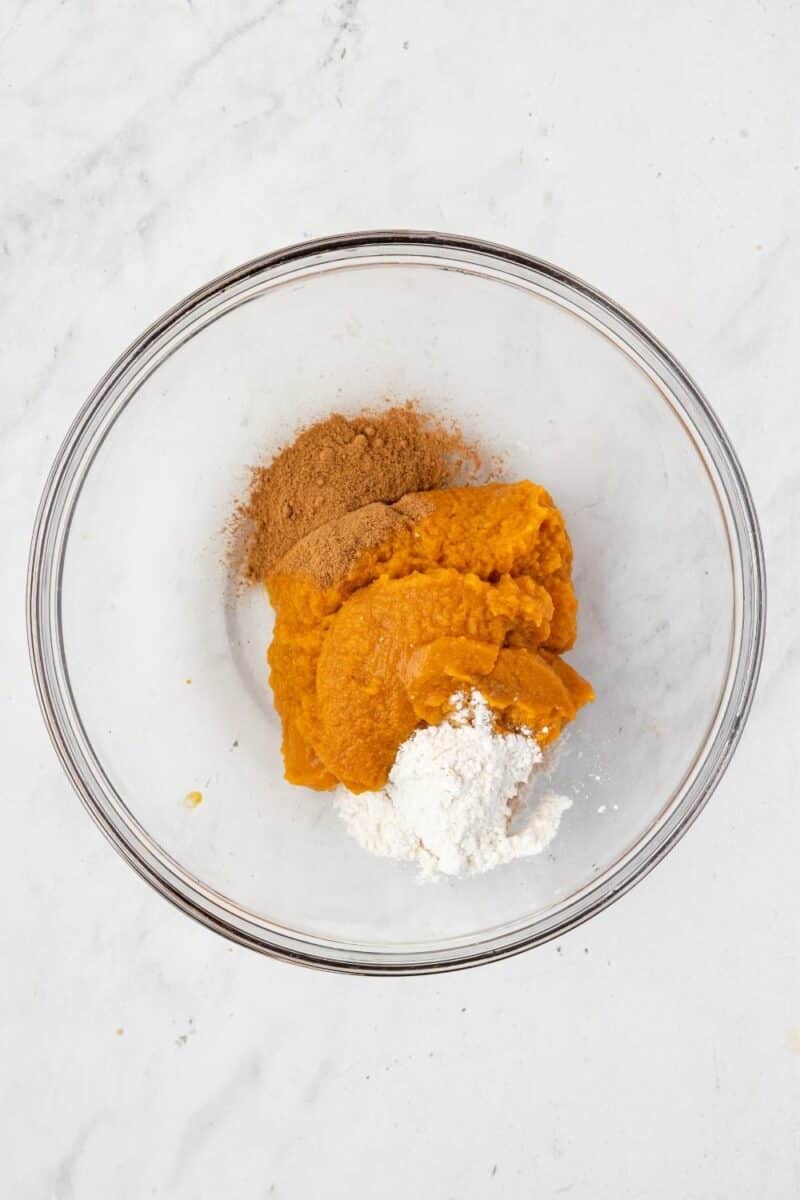 A clear glass bowl contains pumpkin puree, flour, and ground cinnamon on a white marble surface.