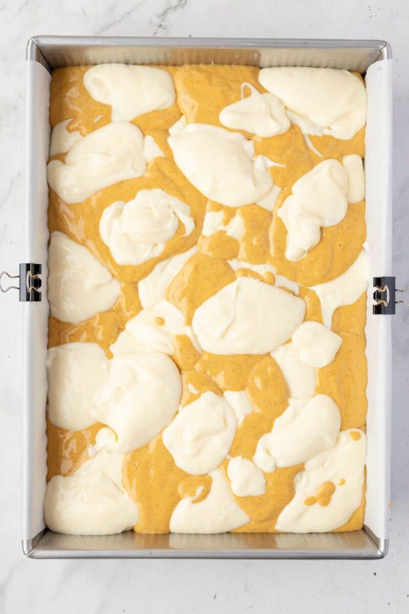 A rectangular metal baking pan lined with parchment paper contains swirled layers of pumpkin and cream cheese batter.