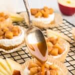 A spoon drizzles caramel sauce over mini caramel apple cheesecakes topped with diced apples, displayed on a cooling rack with apple slices nearby.